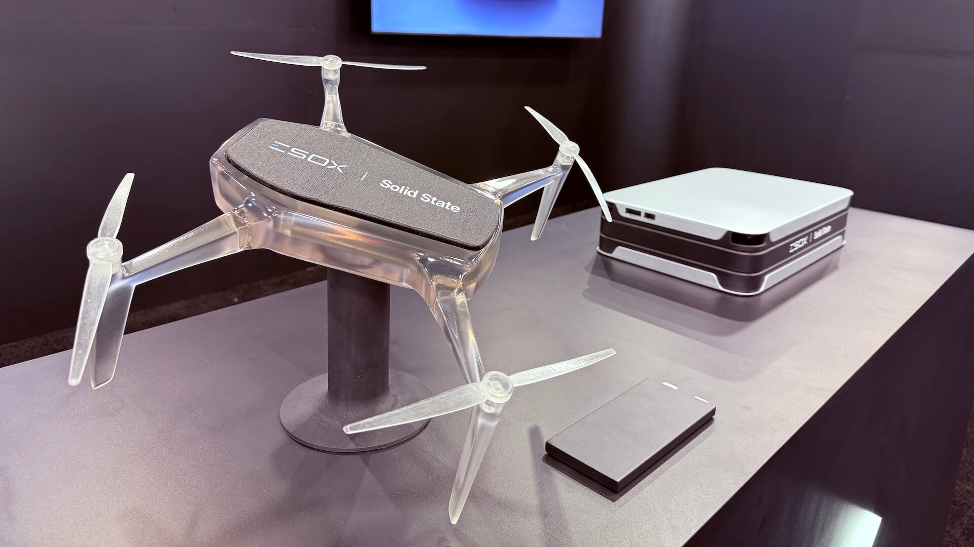 A transparent quadcopter drone with "Solid State" written on top is displayed on a stand, next to a rectangular remote and a compact, stackable electronic device on a table.
